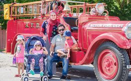 family with three kids posing in front of old red fire truck